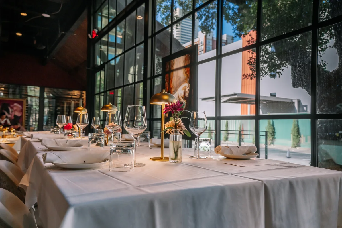 Tables set with white linens, flowers, glassware, and gold lamps along The Kennedy’s windows