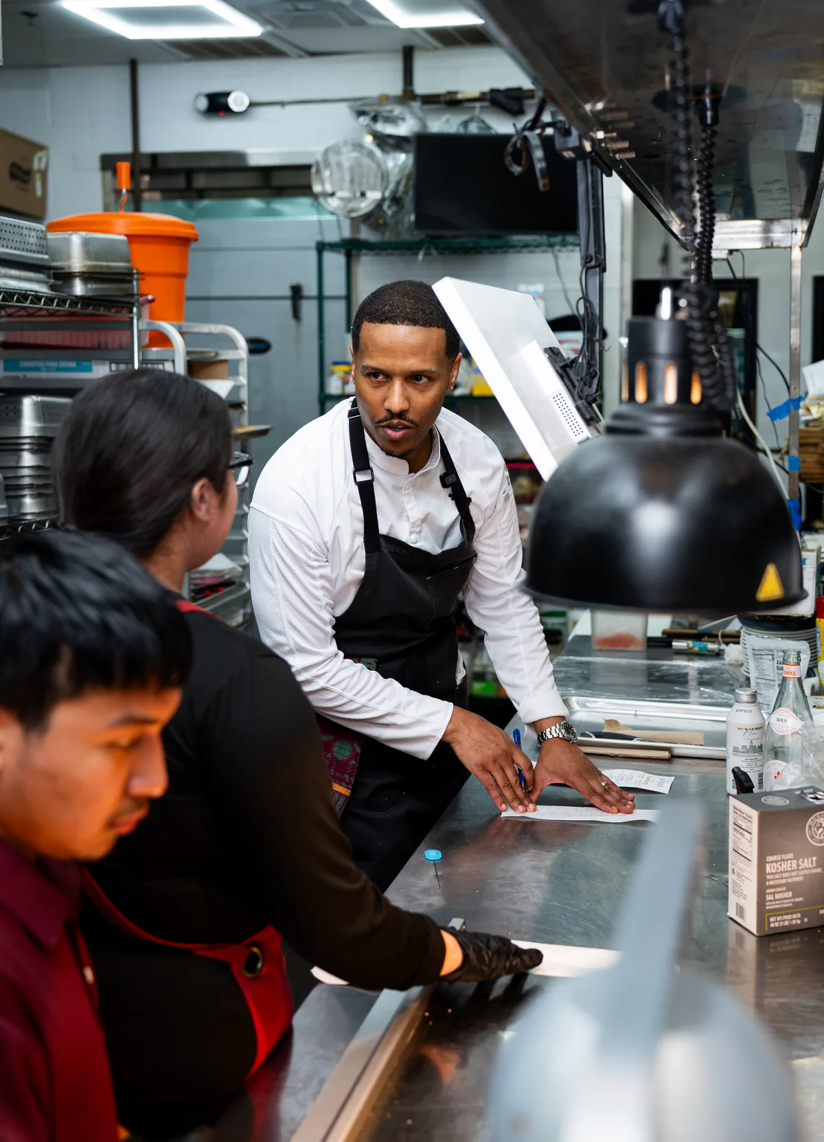Chef in a white jacket and black apron with the kitchen team at the pass
